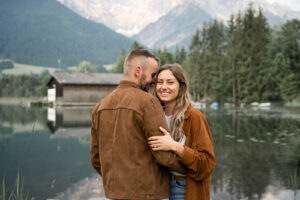 Young couple at Lake Walchsee in Tyrol while the man whispers into the woman’s ear and she looks into the camera smiling happily