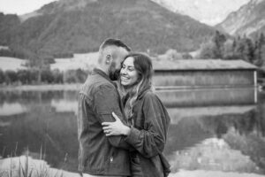 Black-and-white photo of a young couple by a lake, with the man whispering into the woman’s ear while she looks happily into nature. Trees and mountains are reflected in the water.