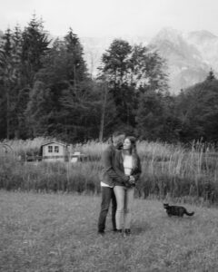 Black-and-white photo of a couple embracing in a field beside a black cat