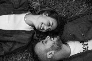 Black-and-white photo of a couple lying on the ground, smiling into each other’s eyes