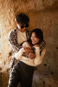Young Korean couple standing in a cave at the Algarve while the man embraces the woman from behind
