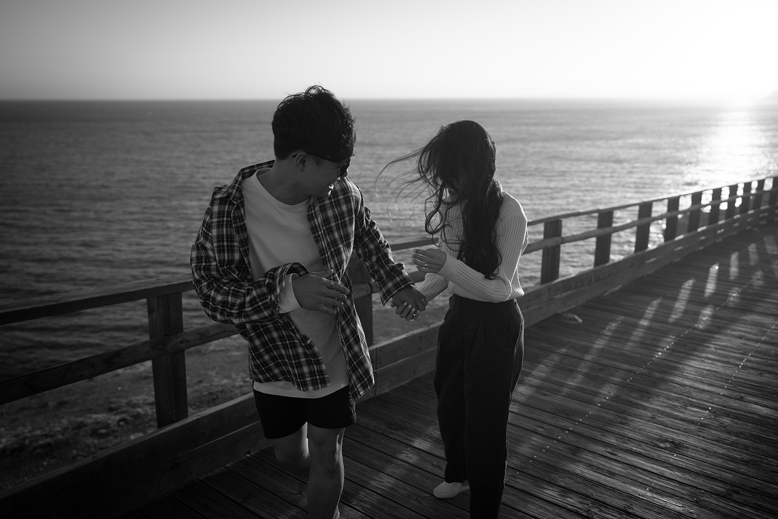 Black-and-white photo of a young couple walking beside the ocean while the wind blows through her hair