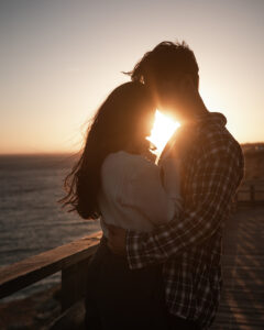 Couple embracing at sunset by the ocean with sunlight shining between their heads