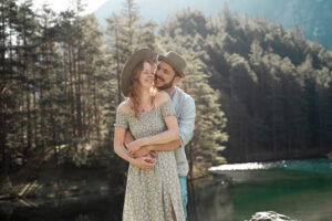 Man embracing his fiancée from behind in backlight while both look over their shoulders laughing