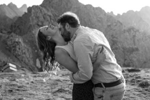 Black-and-white photo of an engaged couple in the mountains while the man kisses his fiancée’s neck