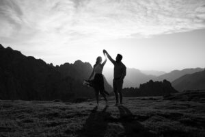 Black-and-white photo of a man twirling his fiancée at sunset on a mountain peak