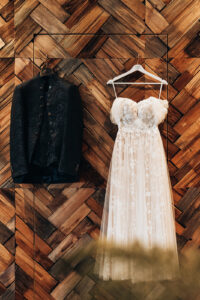 Wedding dress and suit hanging side by side against a herringbone wooden wall