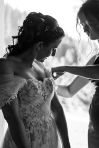 Black-and-white photo of the maid of honor adjusting the sleeves of the wedding dress by a window