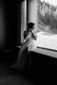 Black-and-white photo of the bride putting on her earrings while sitting by a window