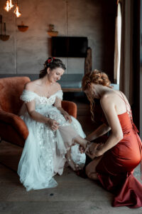The bride sits on a red chair while the maid of honor helps her into her shoes