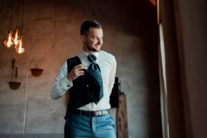 The groom puts on his waistcoat while looking out the window