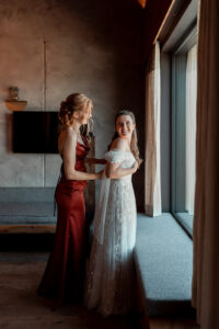 Bride and maid of honor standing by a window, laughing together over their shoulders