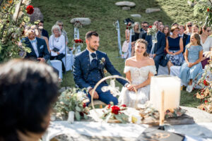 The wedding couple sits hand in hand during the ceremony, smiling ahead