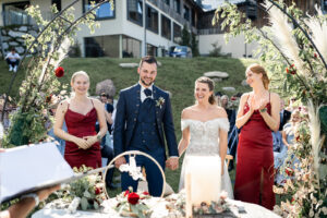 The wedding couple stands hand in hand beside their bridesmaids, smiling