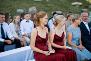 The two bridesmaids sit on a bench, smiling toward the ceremony