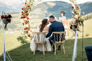 Wedding kiss during the ceremony in front of a circular floral arch with mountain views