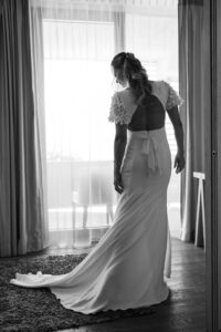 The bride standing in front of a window, photographed from behind, turning to the left while adjusting her dress