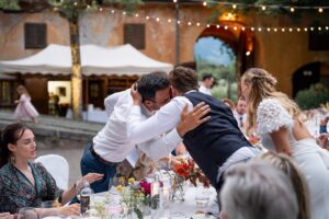 Bride, groom, and wedding attendants sharing a heartfelt hug across the dinner table