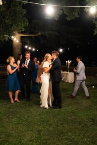 Bride and groom kissing on a field beside their guests beneath string lights