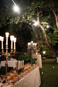 Wedding dessert table with cakes, candles, and string lights with trees in the background