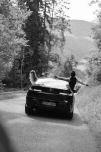 Vintage black-and-white photo of a wedding couple leaning out of a car with their arms spread wide