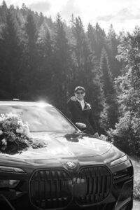 Black-and-white photo of a groom standing beside a car and pointing at the camera