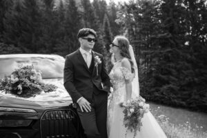 Black-and-white photo of a wedding couple smiling while leaning against their wedding car