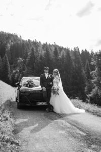 Black-and-white photo of a wedding couple wearing sunglasses beside their wedding car