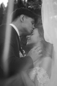 Black-and-white portrait-format photo of a groom kissing his bride on the forehead