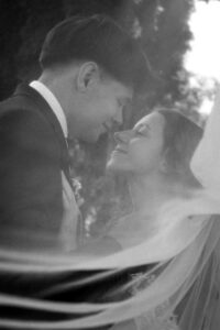 Black-and-white photo beneath the veil of a wedding couple smiling warmly at each other