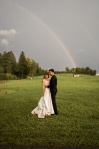 The bride and groom stand on a meadow looking down as the groom embraces his bride, with a rainbow in the background