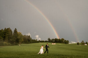 The bride and groom walk hand in hand across a meadow with a full rainbow in the background