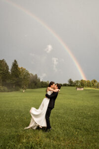 The groom lifts his bride as they kiss in front of a rainbow