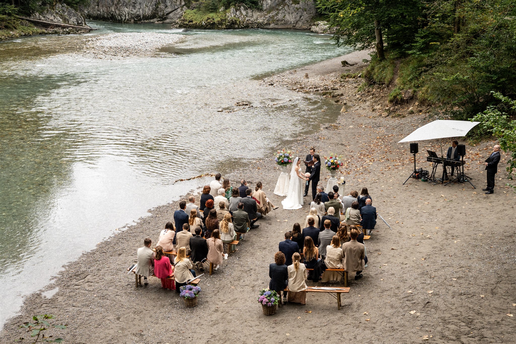 Drone photo of an outdoor wedding ceremony by a riverside with guests