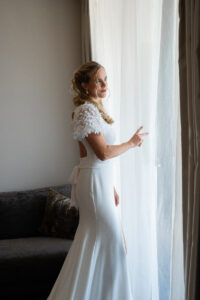 Fully styled bride standing by the window holding back the curtain and looking outside