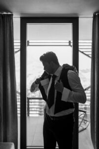 Black and white photo of the groom putting on his vest with help from the best man