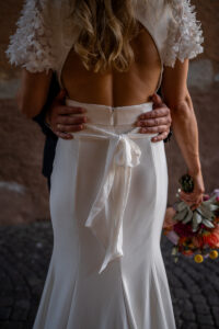 Close-up of the open back of a wedding dress while the groom holds his bride around the waist