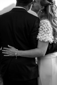 Black and white close-up of a bride hugging her husband from the side and whispering into his ear