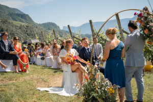 The wedding couple sits among the vineyards in front of their guests, looking toward the celebrant