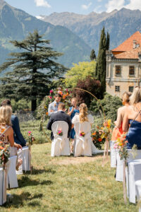 Photographed from behind, the wedding couple sits on white chairs beneath the wedding arch overlooking the mountains and Schloss Rametz