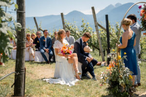 The wedding couple sits among the vineyards during the ceremony while the groom holds their son