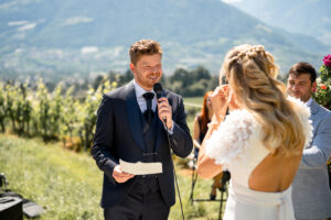Over-the-shoulder view of the groom holding his vows while his wife wipes away tears