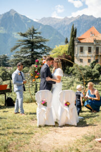 The married couple stands before the wedding arch with their heads together, Schloss Rametz and the mountains behind them