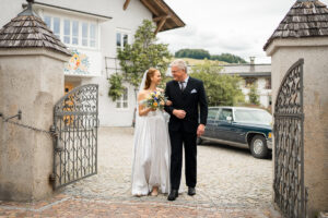 Full-body photo of the bride and her father walking arm in arm toward the church.