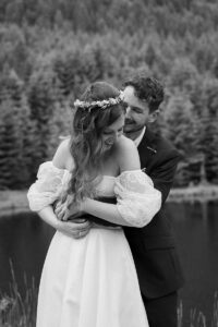 Black-and-white photo of an intimate embrace as the groom hugs his wife from behind and both smile over their shoulders