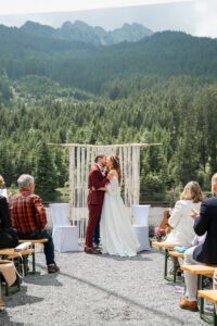 Wedding kiss in front of a macramé ceremony arch with mountains and trees in the background