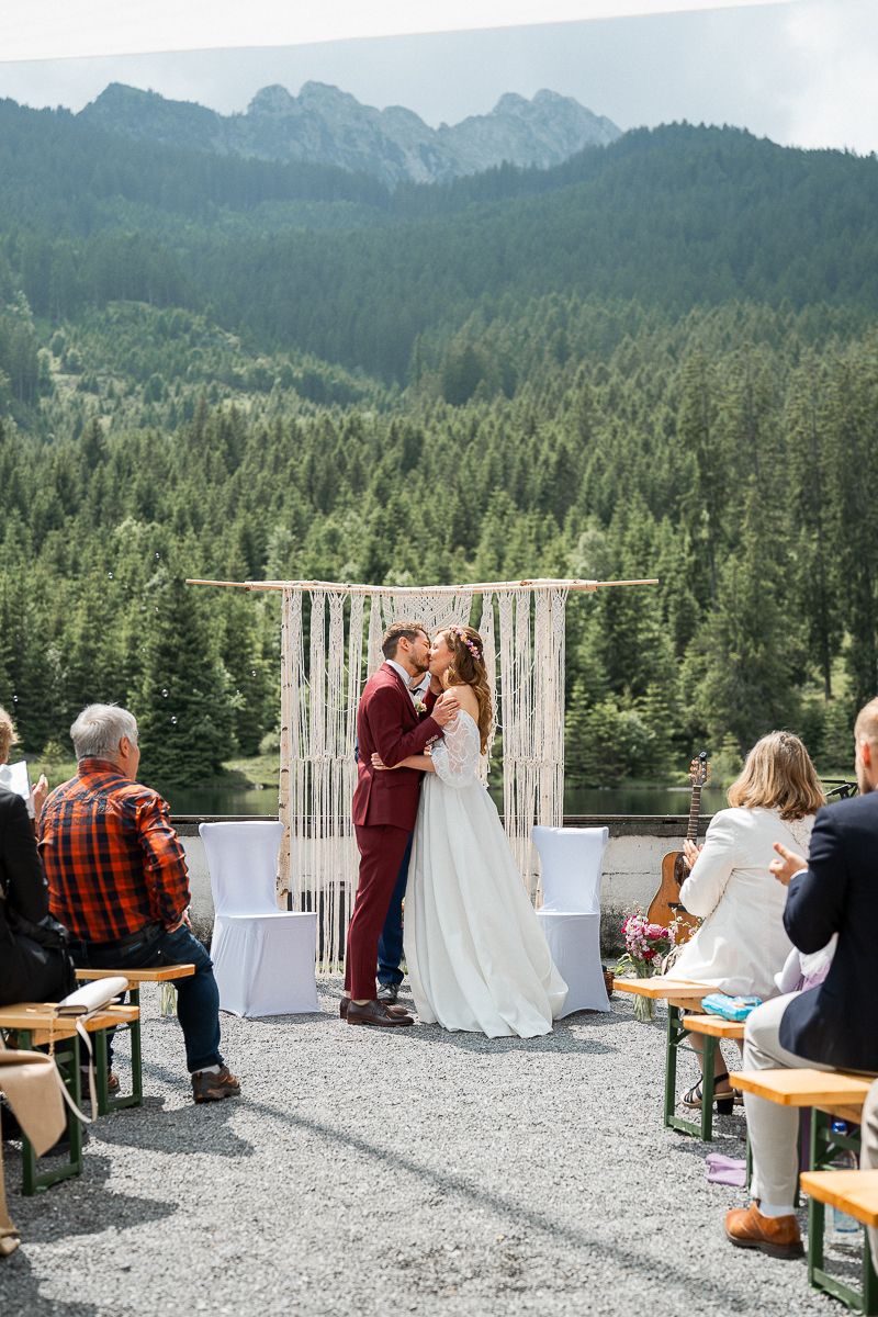Wedding kiss in front of a macramé ceremony arch with mountains and trees in the background