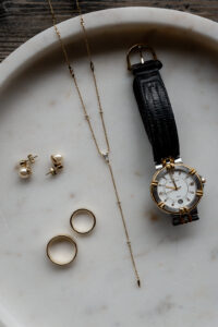 Flat lay of a wristwatch, necklace, earrings, and wedding rings on a marble plate