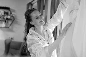 Black-and-white photo of the bride standing by the window looking at her wedding dress