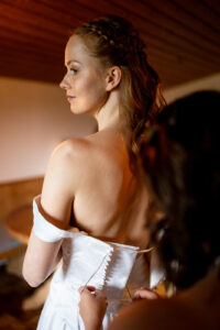High-contrast photo from behind of the bride as her corset is being laced near the window